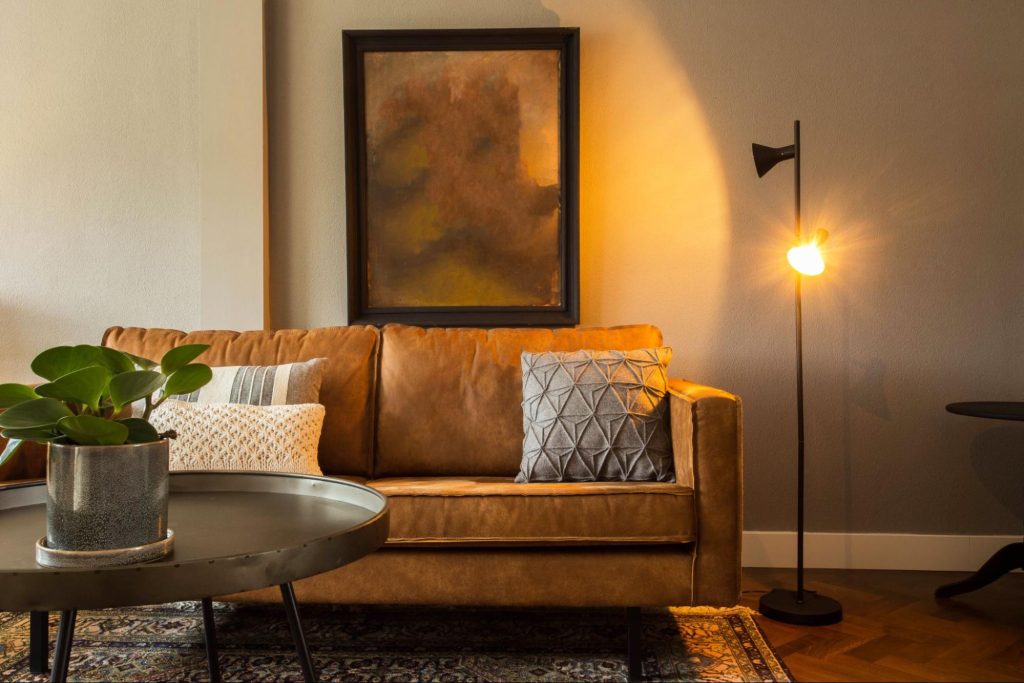 Sala de estar com sofá de couro caramelo, almofadas decorativas, luminária de chão acesa, mesa de centro redonda com planta em vaso e quadro em tons terrosos na parede. Imagem ilustrativa para post sobre tendências de decoração para o outono 2025, do blog da Morar Construtora.